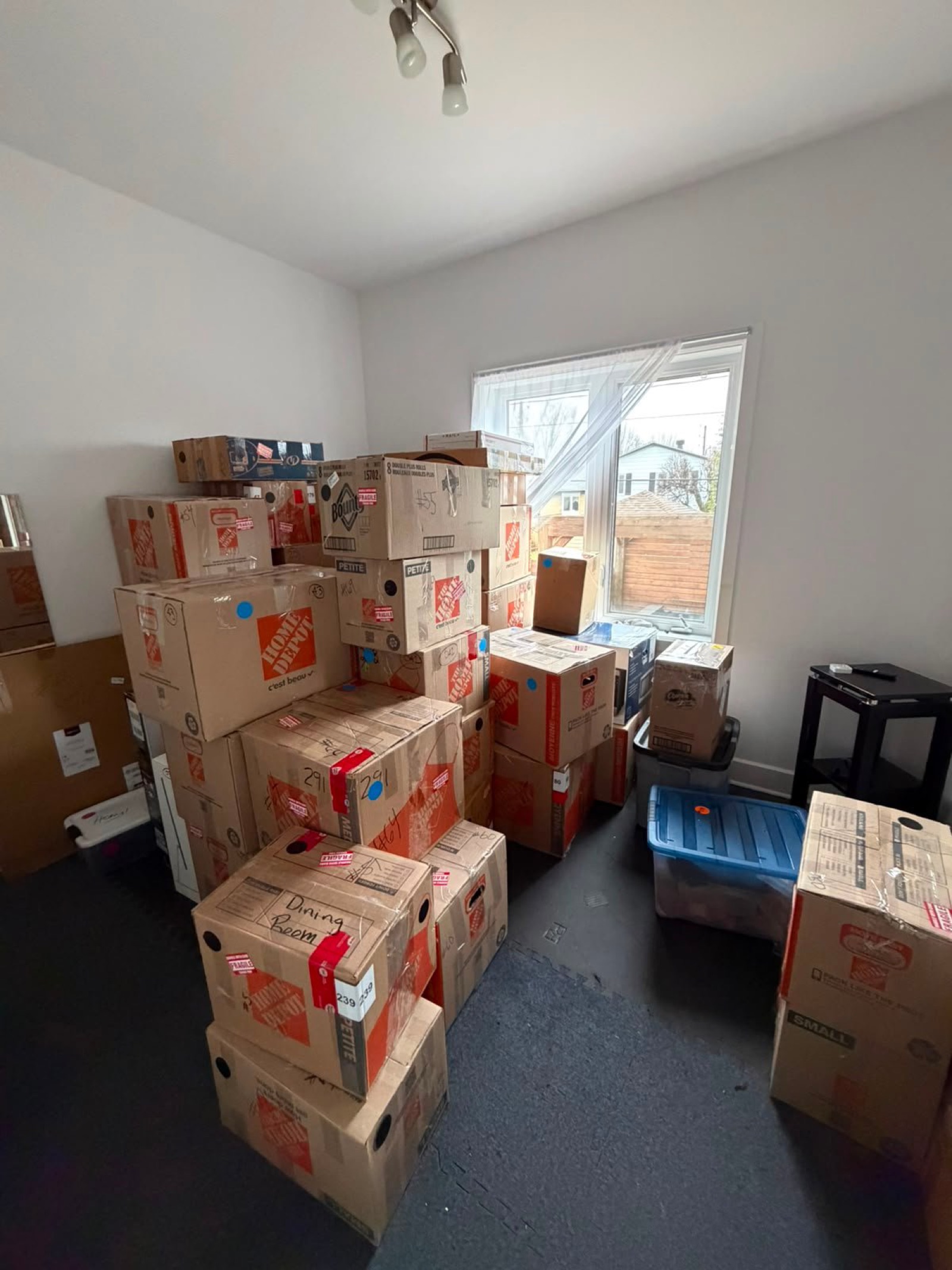 Boîtes de déménagement empilées dans une pièce, avec une fenêtre sur des maisons.